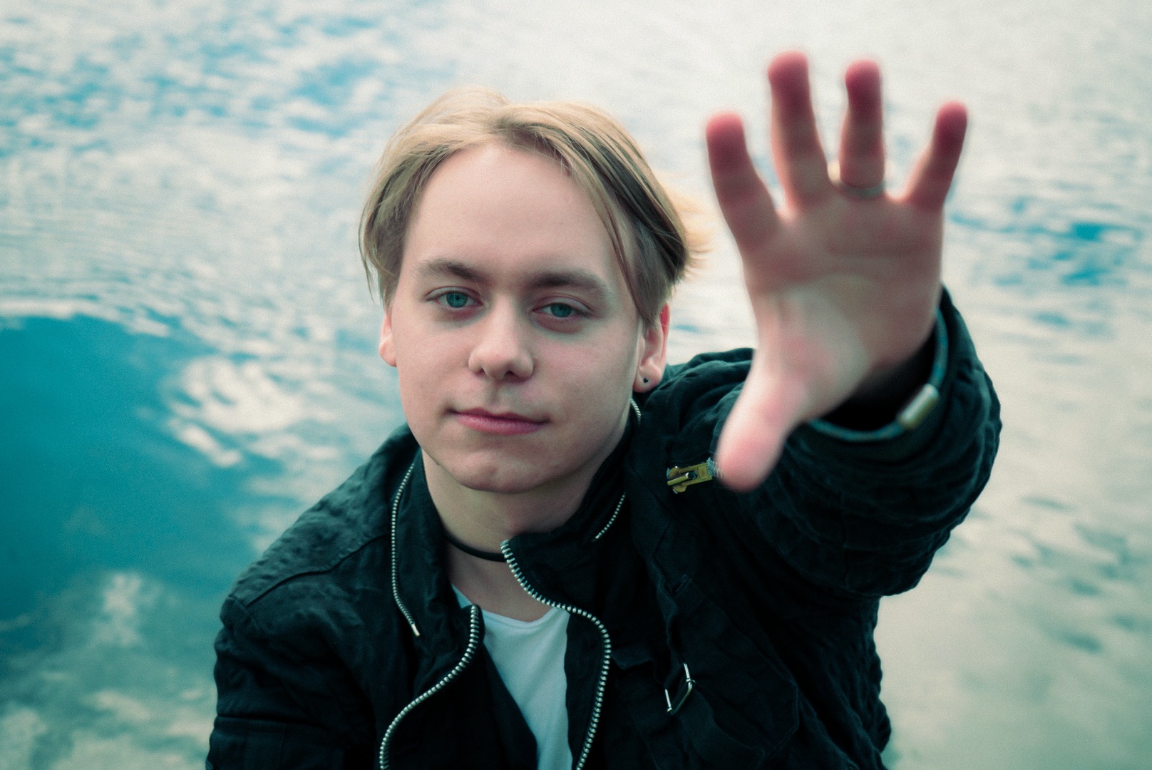 Portret młodego mężczyzny z wyciągniętą dłonią na tle rozmytego, wodnego krajobrazu. Dłoń z pierścionkiem skierowana w stronę obiektywu, tworząca efekt głębi. Mężczyzna ubrany w czarną kurtkę...