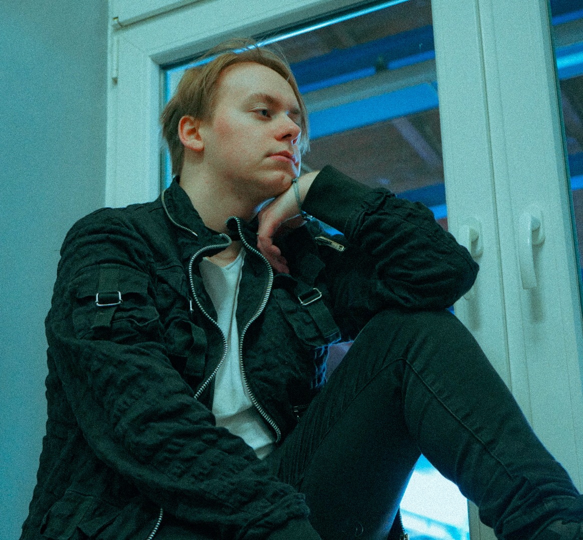 Portret młodego mężczyzny w kurtce siedzącego na parapecie okna w Szczecinie, oświetlonego niebieskim światłem, zamyślony wyraz twarzy.