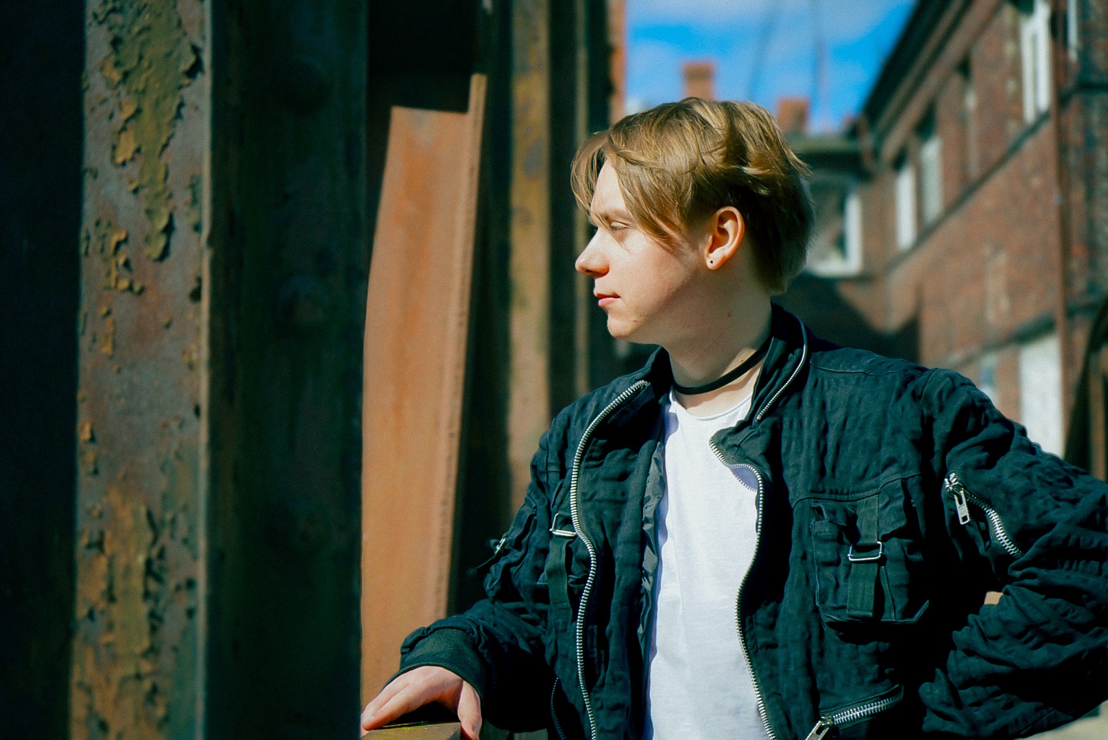 Portret młodego mężczyzny z blond włosami, w czarnej kurtce i chokerze, stojącego na tle ceglanego budynku i metalowych konstrukcji o zróżnicowanej fakturze.