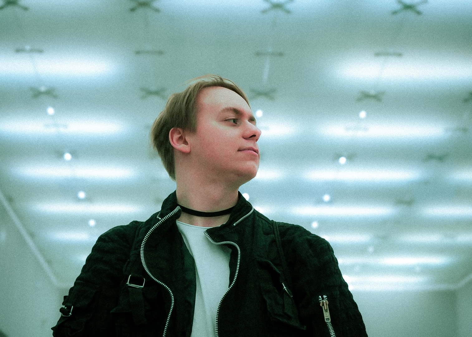 Portret młodego mężczyzny w czarnej kurtce i chokerze, spoglądającego w bok na tle sufitu z geometrycznym wzorem i punktowym oświetleniem.