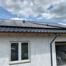 KONKRET Tomasz Augustyniak - Czyszczenie paneli słonecznych na dachu domu jednorodzinnego za pomocą szczotki na teleskopowym kiju, widoczna elewacja budynku w trakcie wykończenia i błękitne niebo z chmurami w tle.