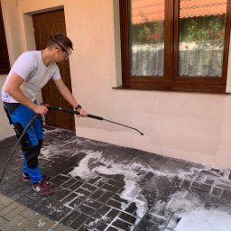 KONKRET Tomasz Augustyniak - Mężczyzna w okularach ochronnych myje kostkę brukową przy pomocy myjki ciśnieniowej, widoczna piana na czyszczonej powierzchni, fragment elewacji budynku z brązowymi drzwiami i oknem w tle.