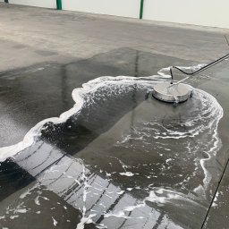 KONKRET Tomasz Augustyniak - Mycie przemysłowej posadzki betonowej za pomocą okrągłej myjki ciśnieniowej, widoczne białe ślady piany i czysta, mokra powierzchnia, pracownik w gumowych butach w prawym rogu.