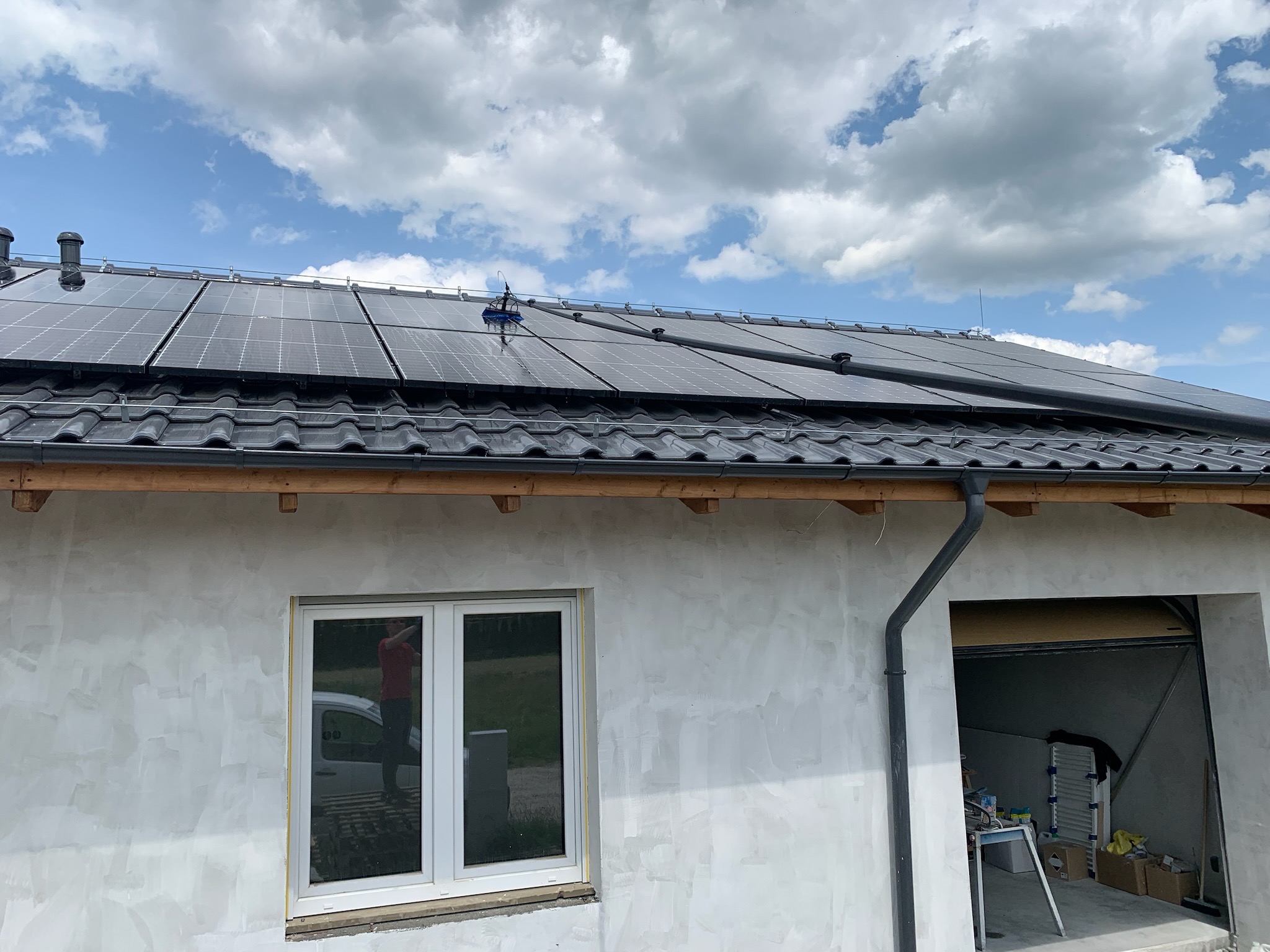 Czyszczenie paneli słonecznych na dachu domu jednorodzinnego za pomocą szczotki na teleskopowym kiju, widoczna elewacja budynku w trakcie wykończenia i błękitne niebo z chmurami w tle.