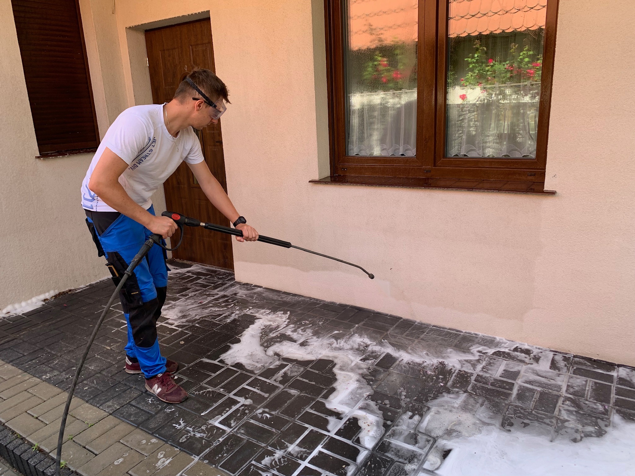 Mężczyzna w okularach ochronnych myje kostkę brukową przy pomocy myjki ciśnieniowej, widoczna piana na czyszczonej powierzchni, fragment elewacji budynku z brązowymi drzwiami i oknem w tle.