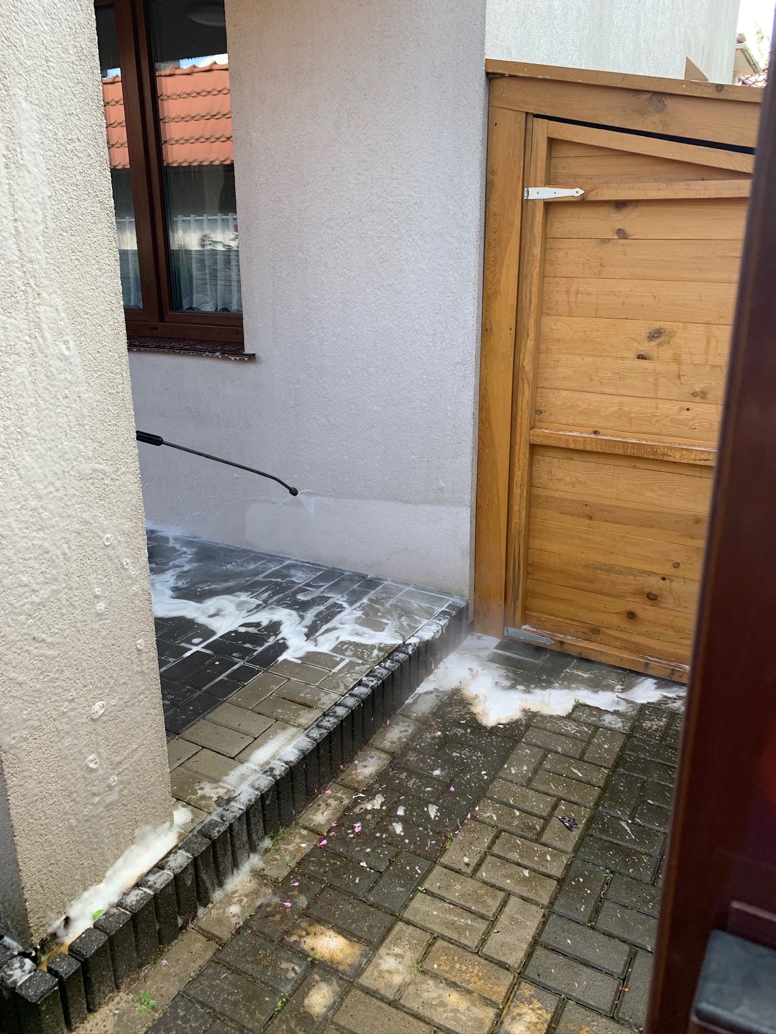 Czyszczenie kostki brukowej podwórza z użyciem myjki ciśnieniowej, widoczne piana i mokra powierzchnia, drewniana brama w tle.