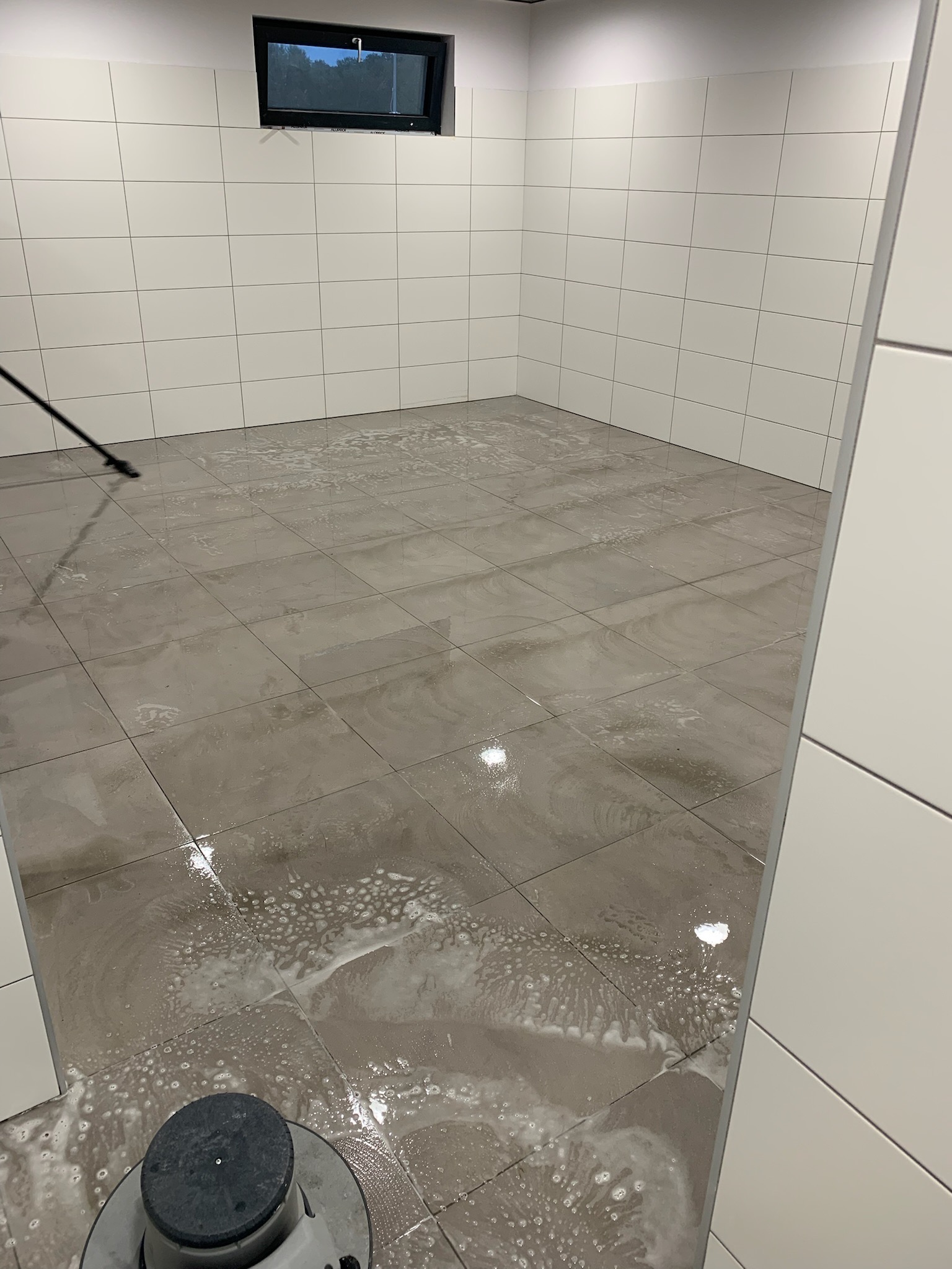 Szorowarka do podłóg w trakcie pracy, pozostawiająca mokre ślady i pianę na szarych, kwadratowych płytkach w pomieszczeniu z białymi, kafelkowymi ścianami i małym oknem pod sufitem.