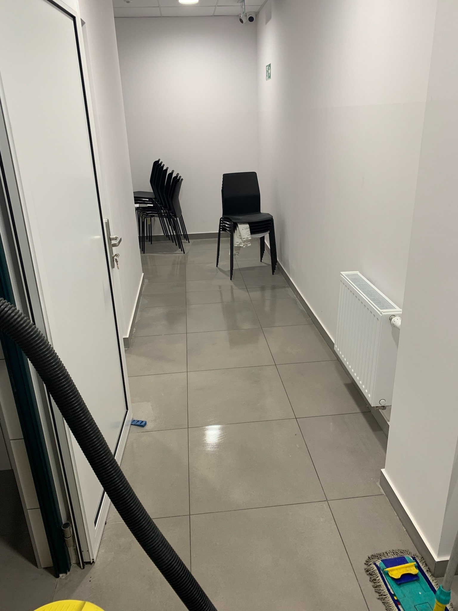 Korytarz biurowy z ułożonymi krzesłami, grzejnikiem i mopem, w trakcie sprzątania lub po sprzątaniu, widoczny wąż odkurzacza.