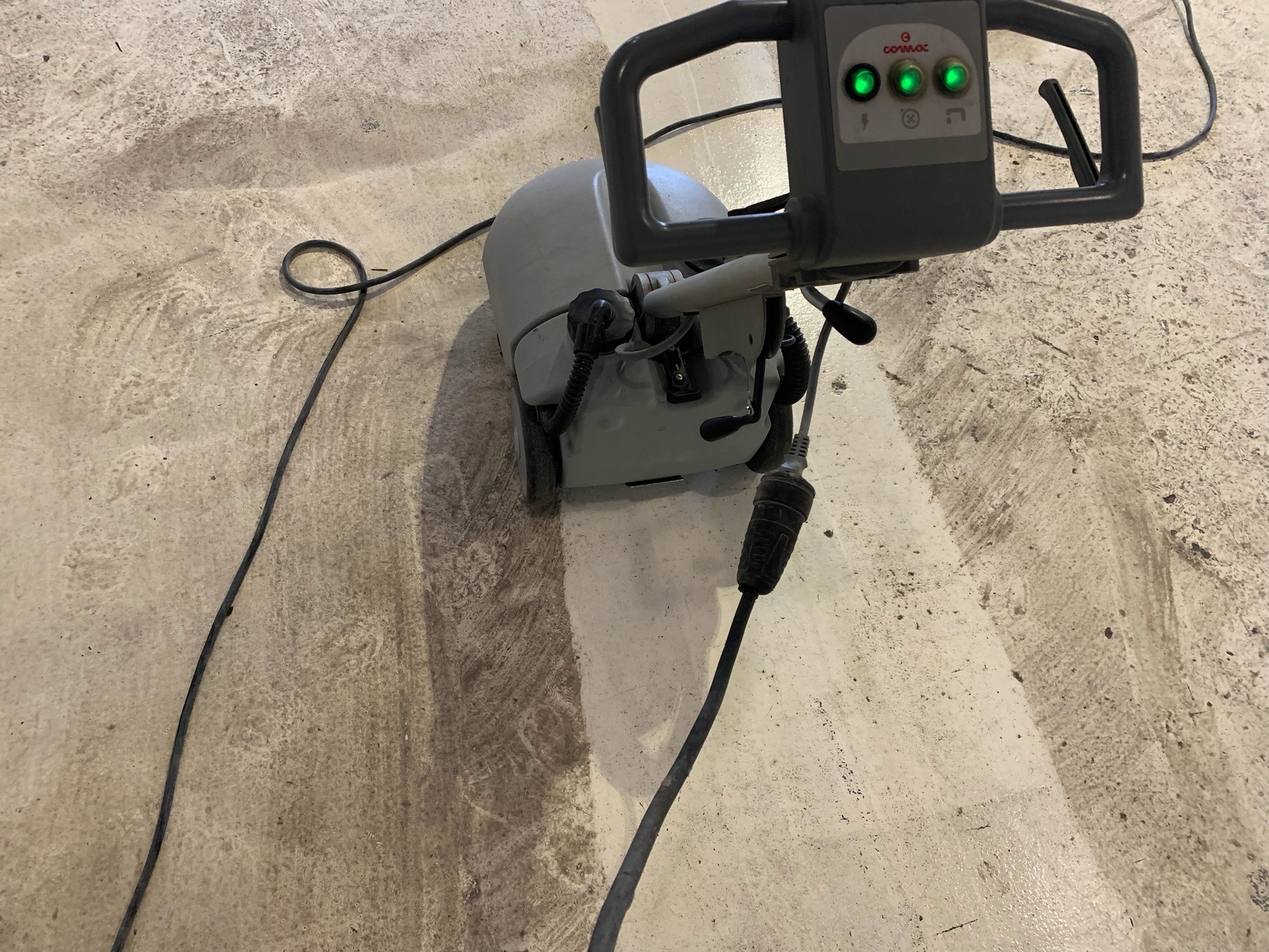 Profesjonalna maszyna czyszcząca Comac z trzema zielonymi diodami, pracująca na betonowej posadzce, widoczny efekt czyszczenia.