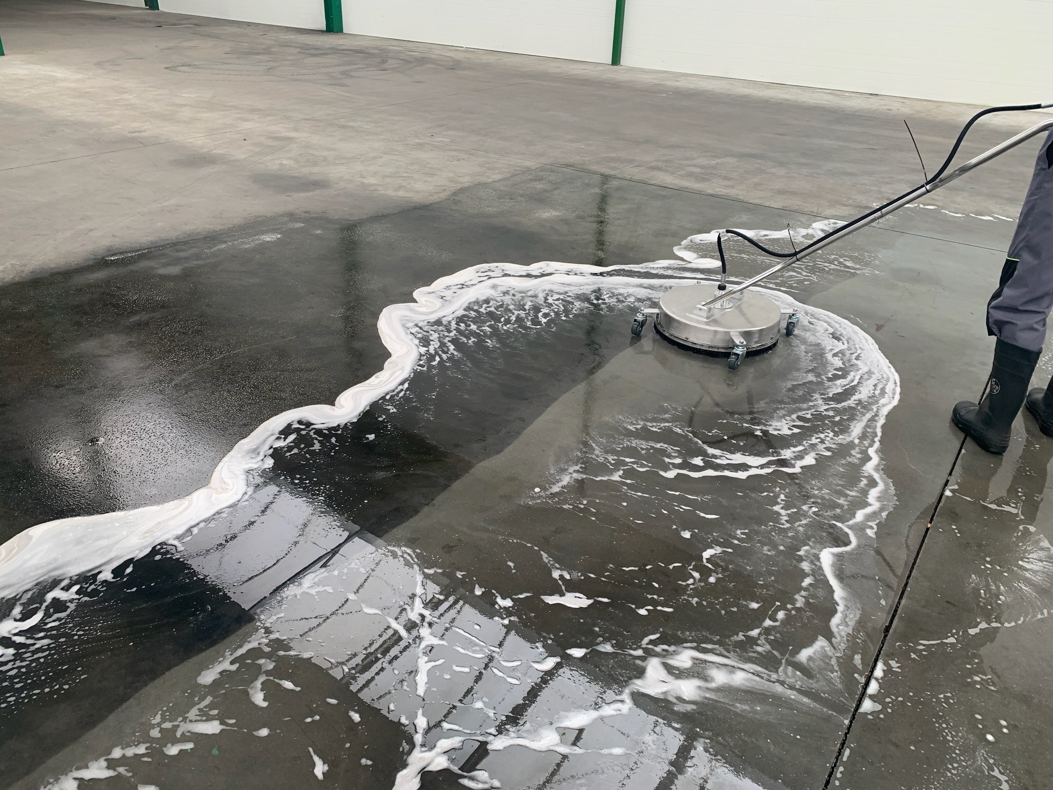 Mycie przemysłowej posadzki betonowej za pomocą okrągłej myjki ciśnieniowej, widoczne białe ślady piany i czysta, mokra powierzchnia, pracownik w gumowych butach w prawym rogu.