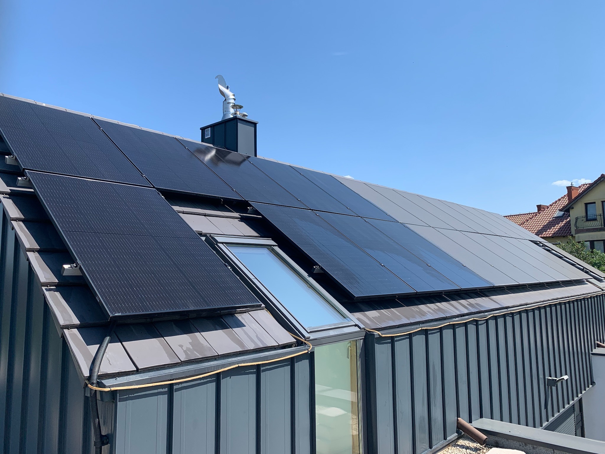 Ciemnoszare panele fotowoltaiczne częściowo zamontowane na dachu z szarej blachy trapezowej, widoczne okno dachowe i ozdobny wiatrowskaz w kształcie koguta na kominie.