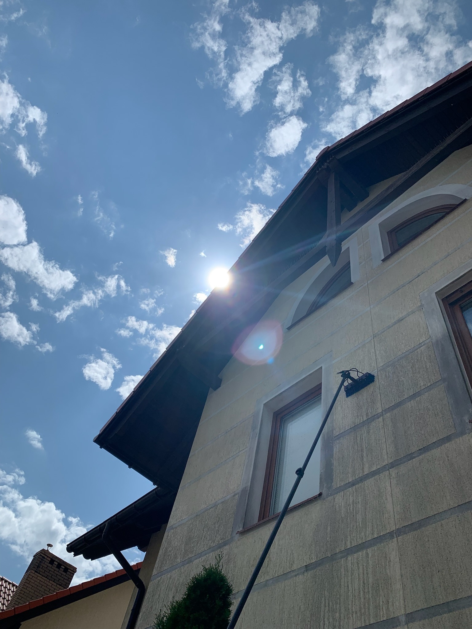 Mycie elewacji domu jednorodzinnego przy użyciu szczotki na teleskopowym wysięgniku, widoczne smugi czyszczące, jasne niebo z chmurami w tle.