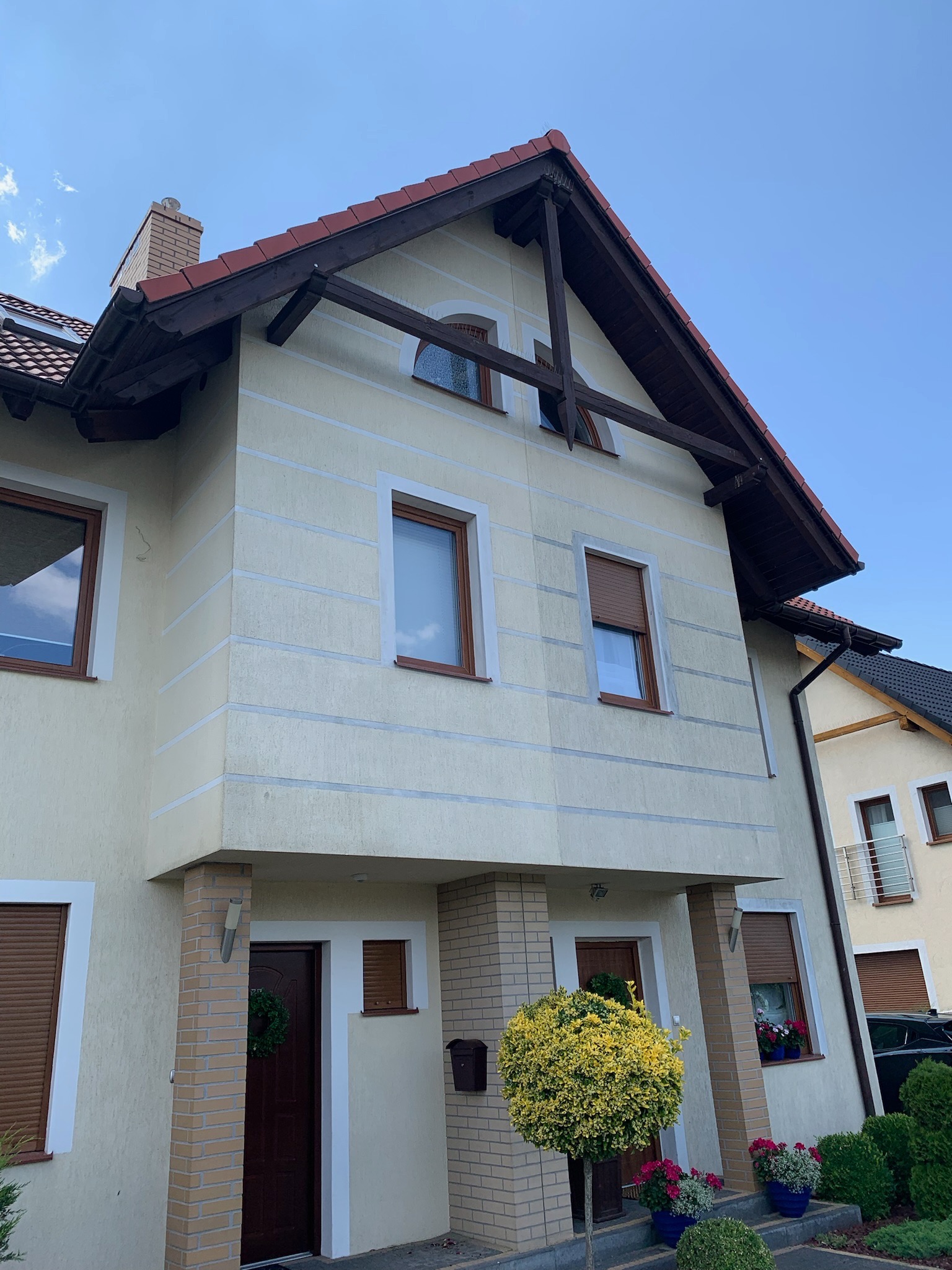 Dwupiętrowy dom bliźniak z beżową elewacją, brązowymi oknami i dachem z czerwonej dachówki, z widocznym fragmentem ogrodu z ozdobnymi krzewami i kwiatami w donicach, sfotografowany w słoneczny dzień.