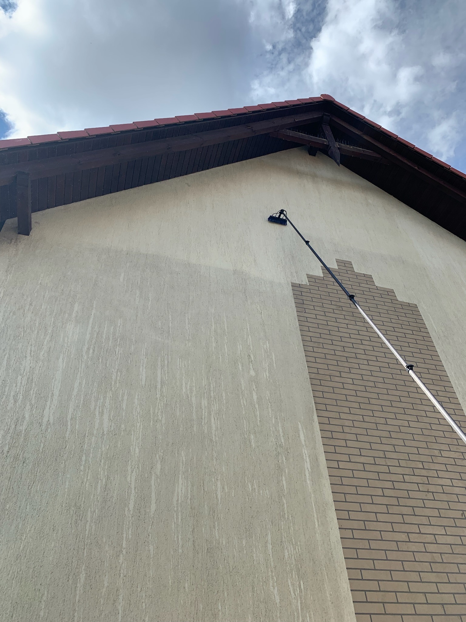 Elewacja budynku z dekoracyjnym wzorem cegieł czyszczona teleskopową szczotką pod kątem, widoczne smugi po czyszczeniu i fragment dachu z czerwonej dachówki.