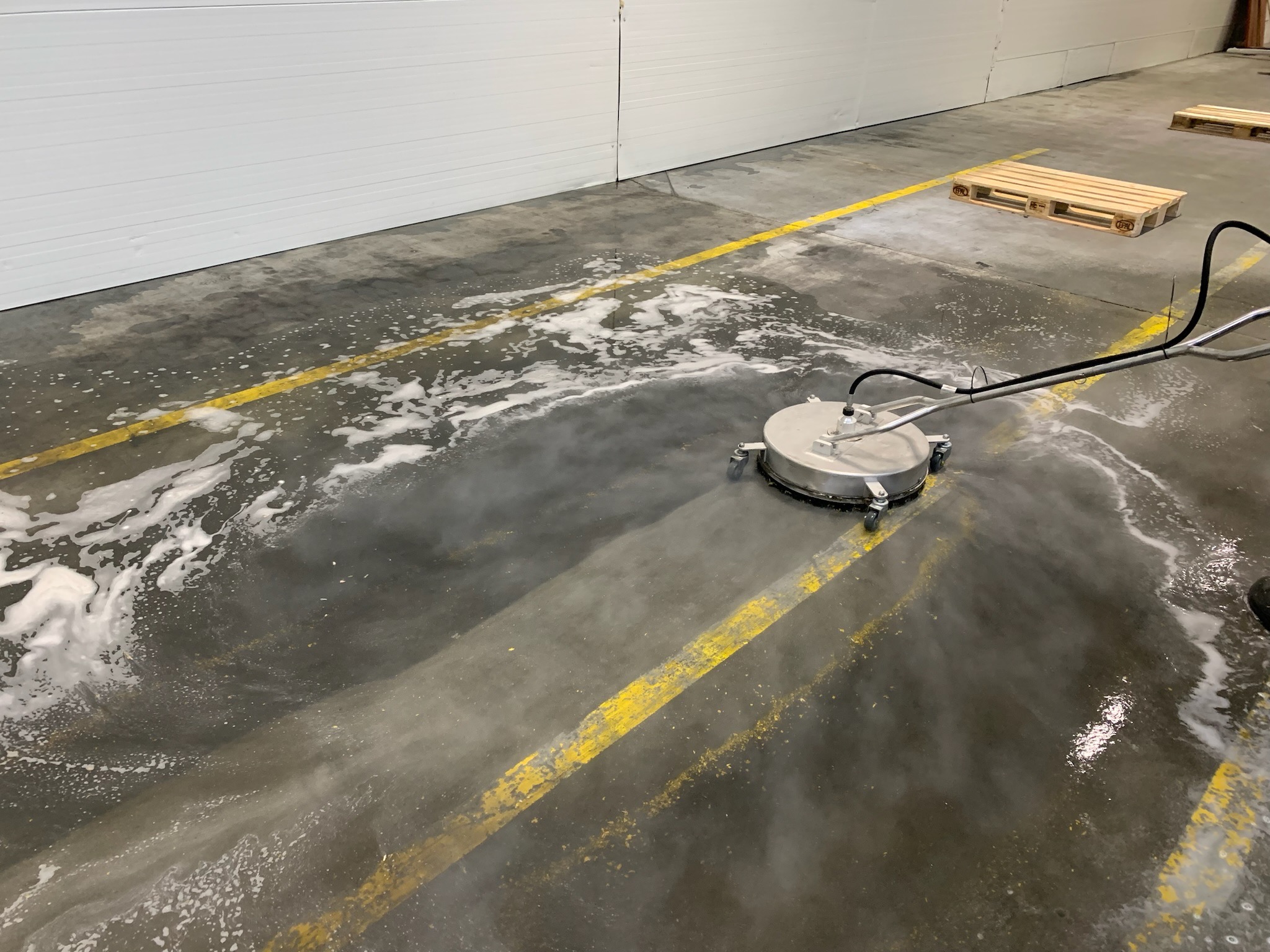 Profesjonalne urządzenie do czyszczenia posadzek przemysłowych w trakcie pracy, widoczne ślady piany i pary wodnej na betonowej podłodze z żółtymi liniami, w tle palety.