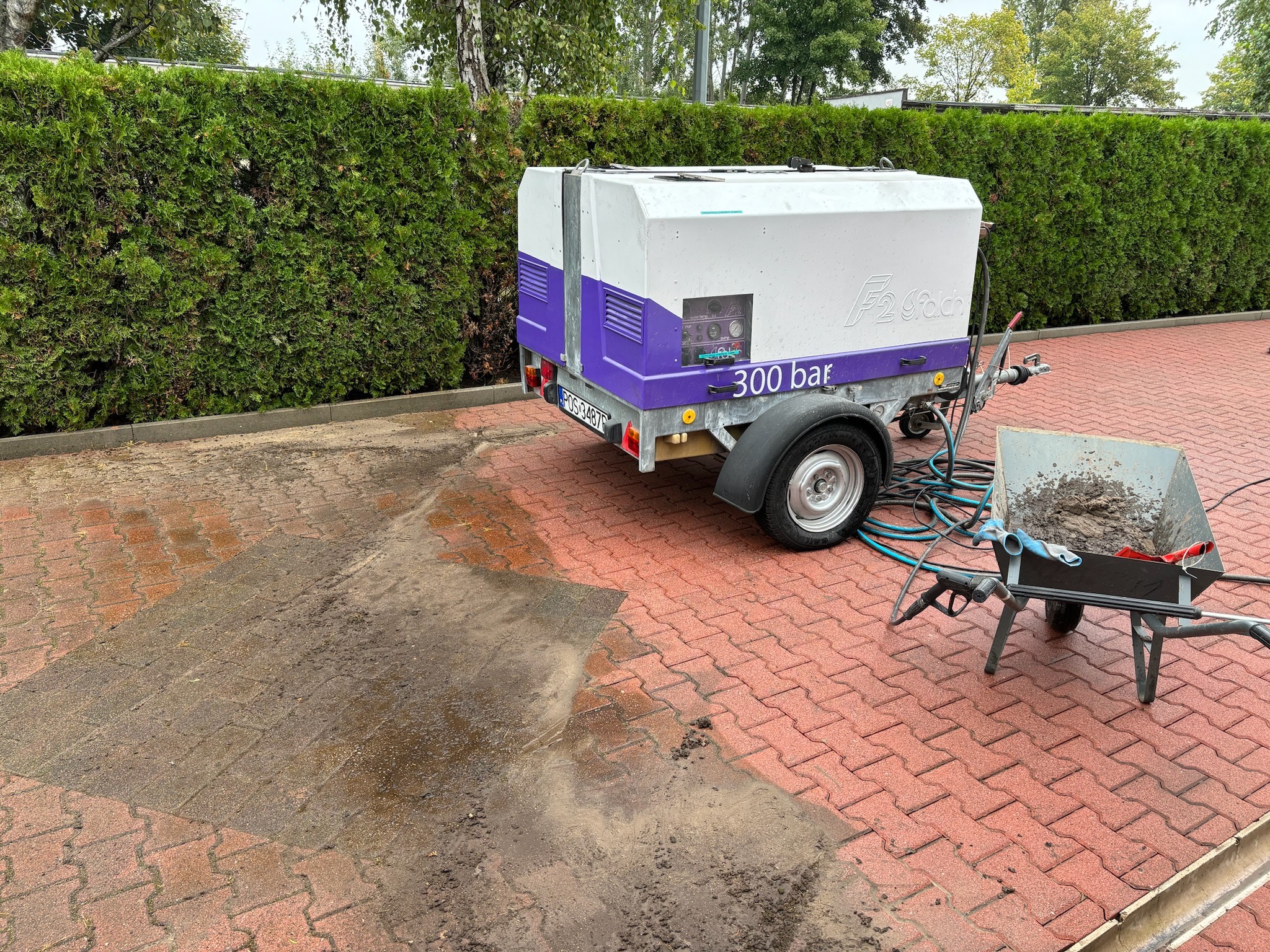 Przyczepa z myjką ciśnieniową F2 Fach 300 bar na tle zielonego żywopłotu i czerwonej kostki brukowej, widoczny fragment umytej kostki oraz taczka z zabrudzeniami.