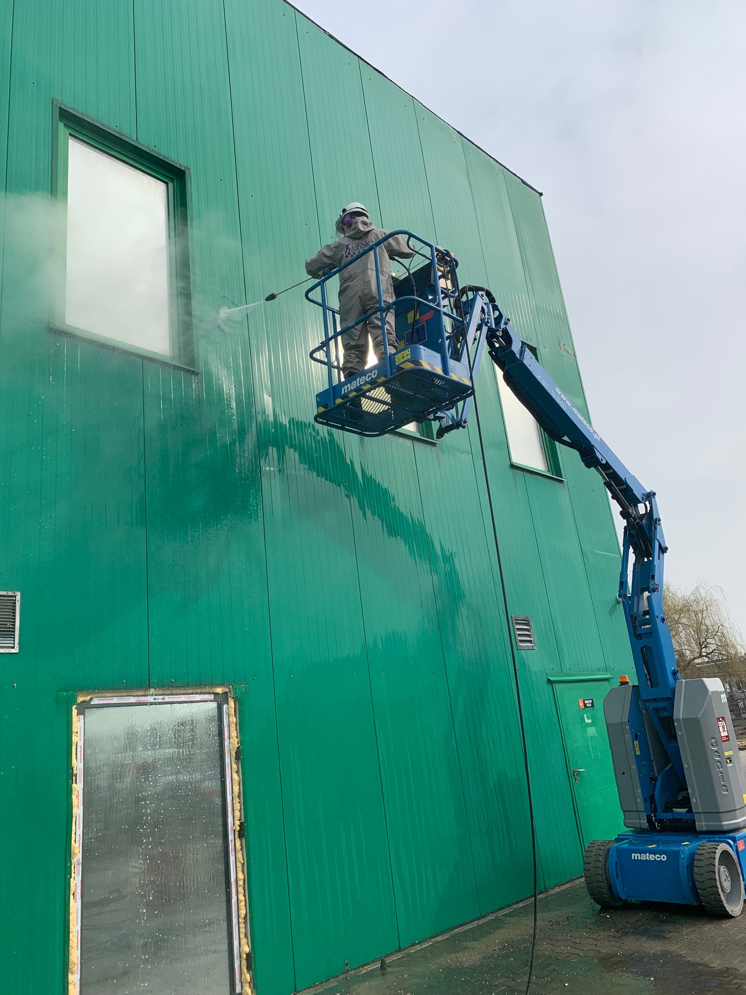 Pracownik w kombinezonie ochronnym na podnośniku koszowym czyści zieloną elewację budynku myjką ciśnieniową w pobliżu okna.