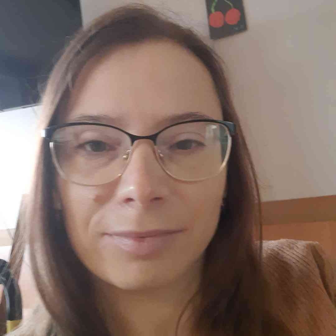 Portret kobiety w okularach o prostokątnych oprawkach, uśmiechającej się do kamery, na tle ściany z dekoracją w kształcie czereśni.