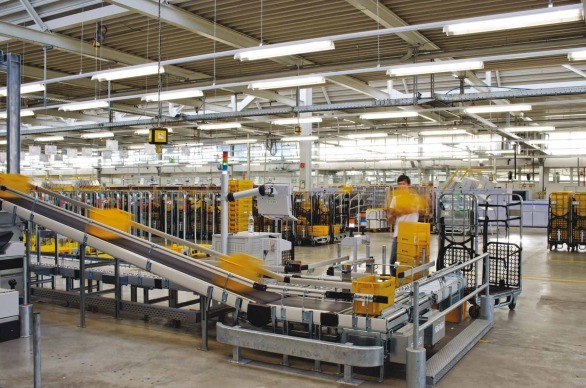 Fluorescencyjne lampy oświetlają halę magazynową z taśmociągiem transportującym żółte pojemniki, widoczny pracownik w tle.