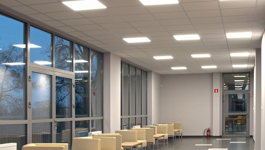 Hallway with suspended ceiling and evenly spaced square LED panel lights, large windows reflecting trees, and waiting area seating.