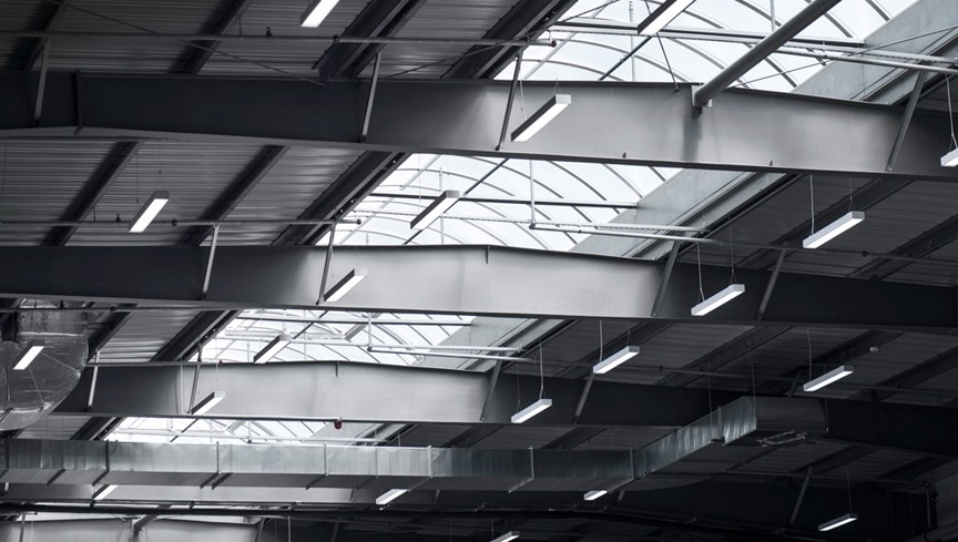 Widok na sufit hali przemysłowej z widocznymi lampami LED, kanałami wentylacyjnymi i świetlikami dachowymi.