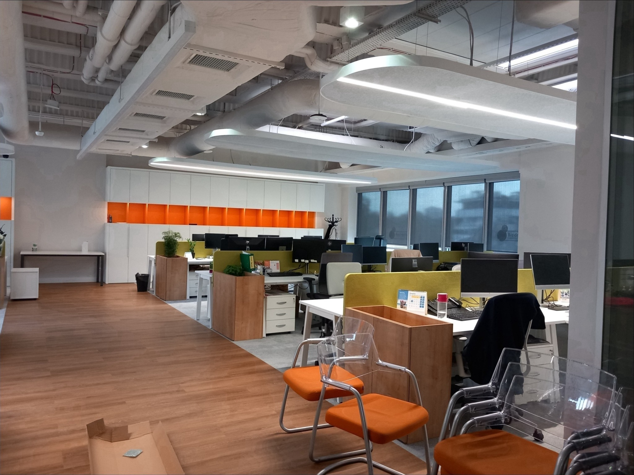 Nowoczesne biuro z otwartą przestrzenią, widoczne biurka z komputerami, pomarańczowe krzesła i podwieszane, linearne oprawy oświetleniowe LED.