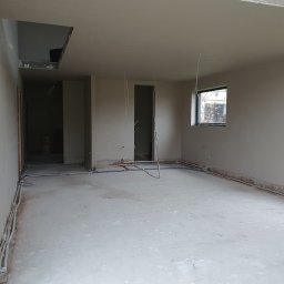 Seel Tynk - Wnętrze w trakcie prac wykończeniowych: gładkie ściany, instalacje w podłodze, surowy betonowy strop z oknem dachowym. Widoczne przewody i rury instalacyjne.