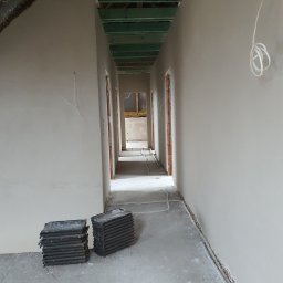 Seel Tynk - Korytarz w surowym stanie po nałożeniu tynków. Widoczne ściany w jasnym odcieniu szarości, surowy beton na podłodze i elementy instalacji elektrycznej.