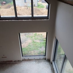 Seel Tynk - Wnętrze domu w trakcie prac wykończeniowych: ściany po nałożeniu gładzi, duże okna z widokiem na zewnątrz, surowa podłoga. Widok z góry.