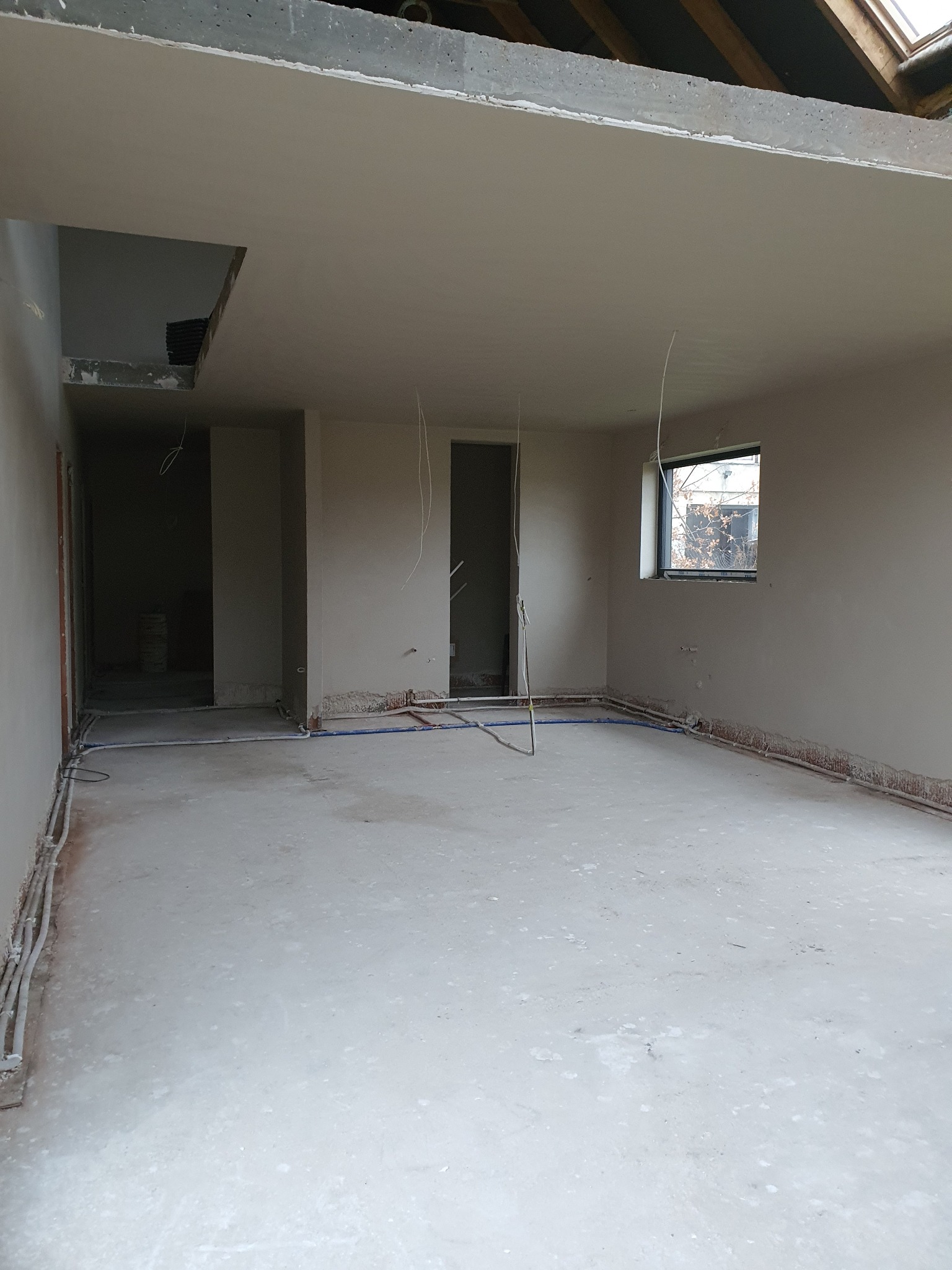 Wnętrze w trakcie prac wykończeniowych: gładkie ściany, instalacje w podłodze, surowy betonowy strop z oknem dachowym. Widoczne przewody i rury instalacyjne.