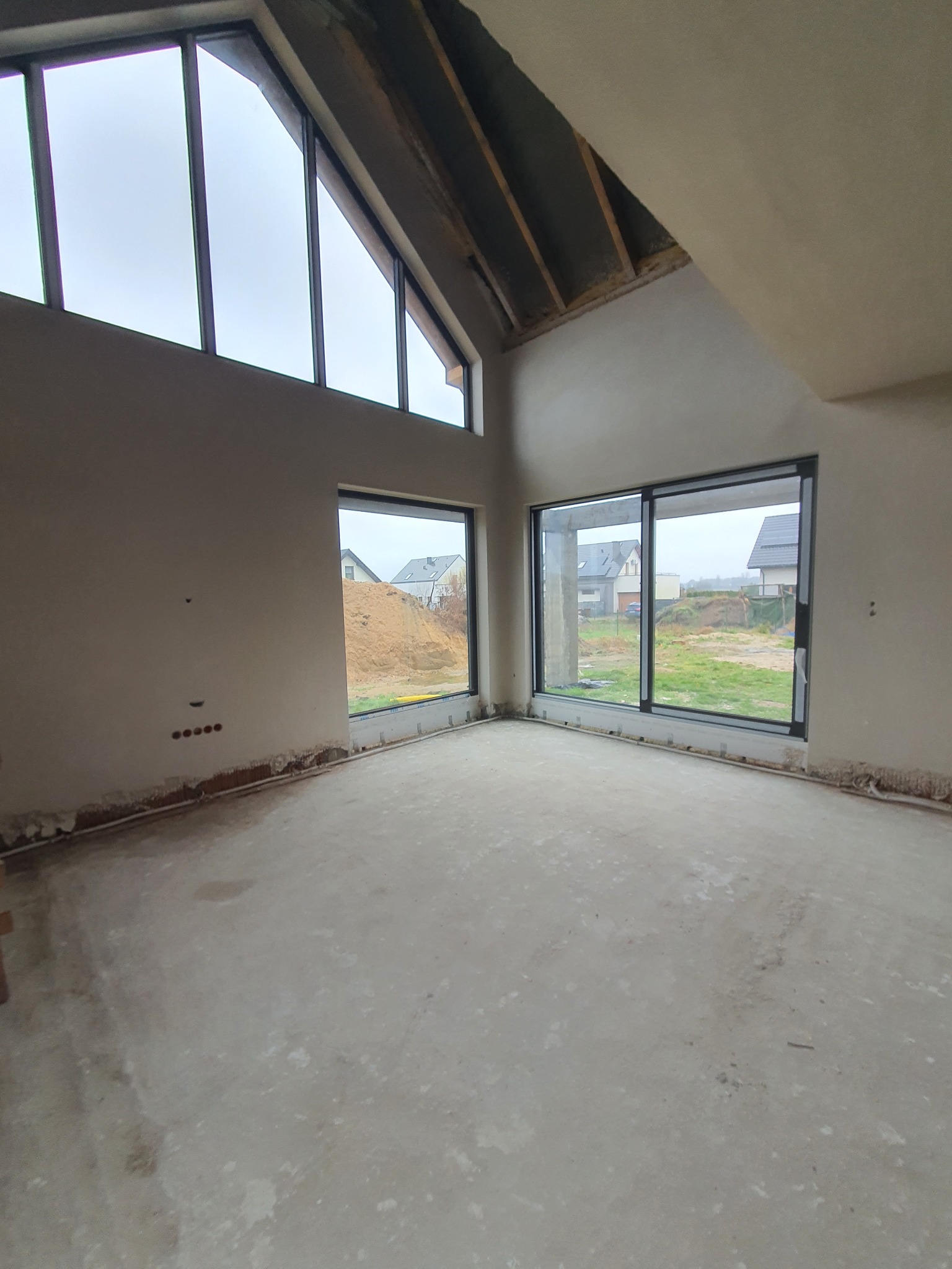 Surowe wnętrze z dużymi oknami i wysokim stropem, ściany przygotowane pod malowanie, widoczne instalacje elektryczne. Wnętrze w stanie deweloperskim.