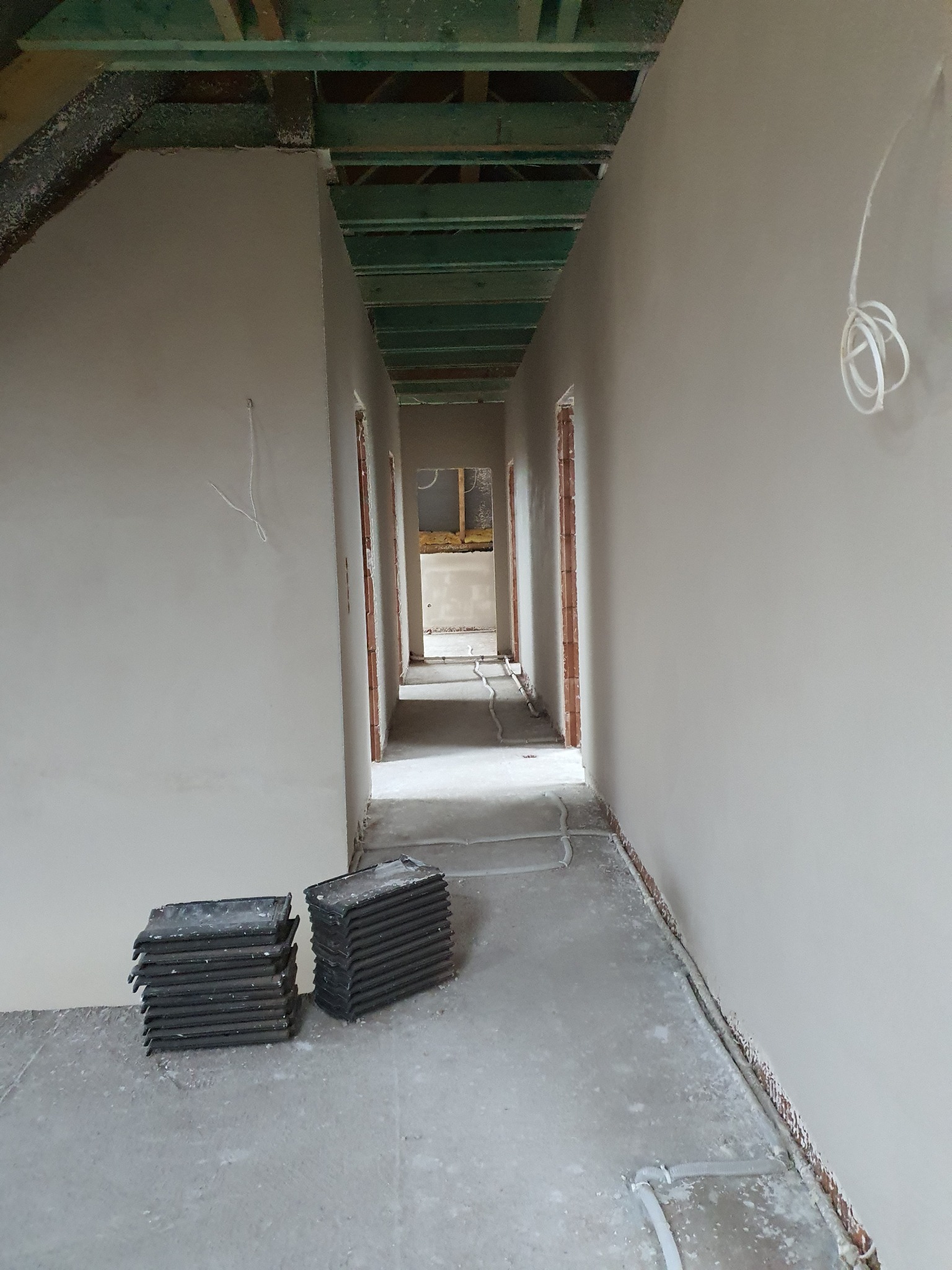 Korytarz w surowym stanie po nałożeniu tynków. Widoczne ściany w jasnym odcieniu szarości, surowy beton na podłodze i elementy instalacji elektrycznej.
