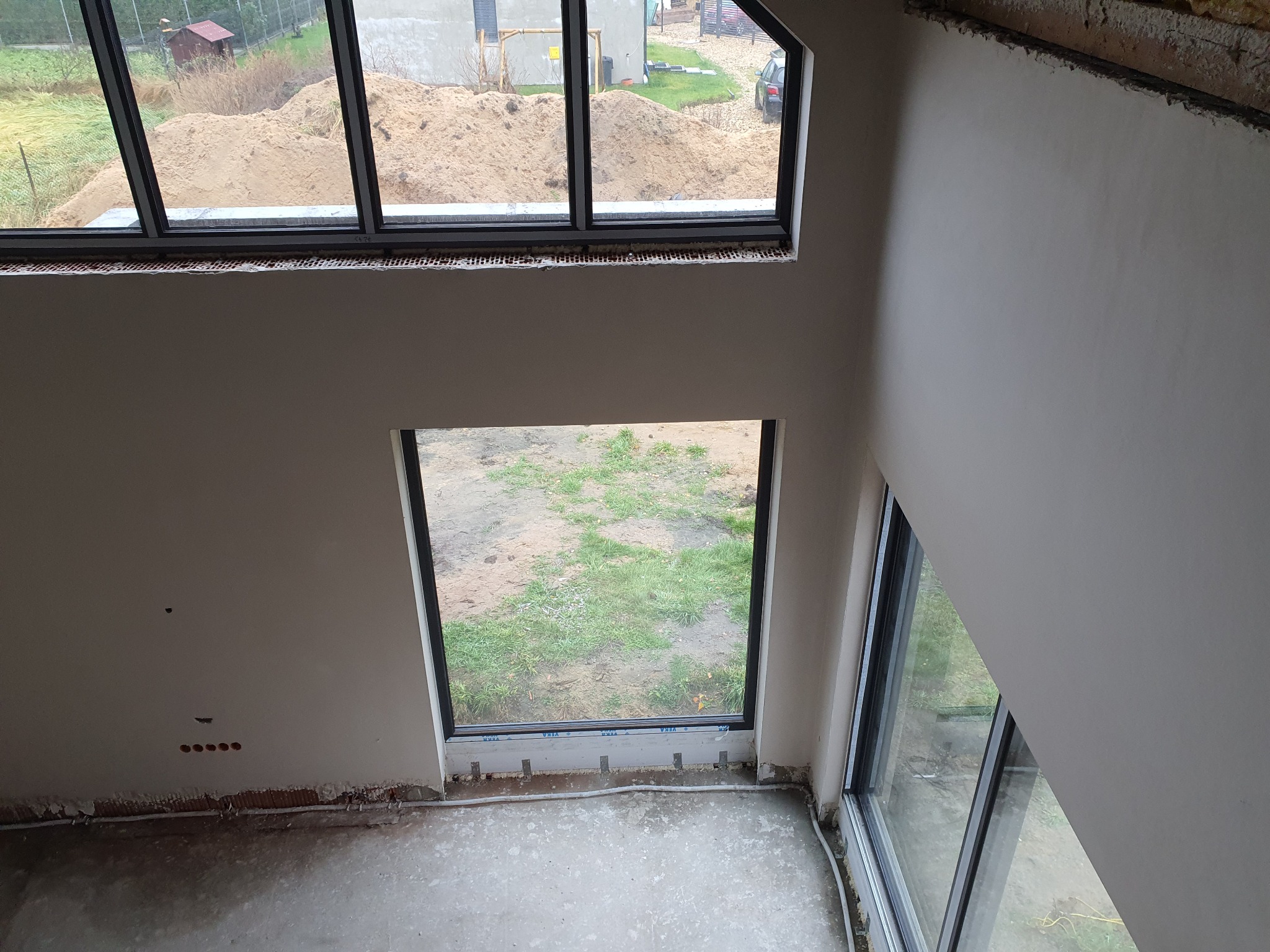 Wnętrze domu w trakcie prac wykończeniowych: ściany po nałożeniu gładzi, duże okna z widokiem na zewnątrz, surowa podłoga. Widok z góry.