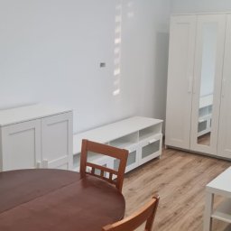 Pucuś Bert - Jasne wnętrze pokoju po odświeżeniu: białe ściany, meble, drewniana podłoga i okrągły stół z krzesłami. Minimalistyczny styl wykończenia.