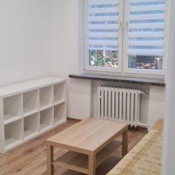 Pucuś Bert - Jasne wnętrze pokoju po odświeżeniu ścian, z nową podłogą i meblami. Widoczne białe ściany, okno z roletami i grzejnik.