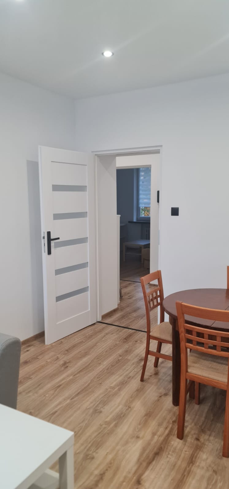 Jasne wnętrze po remoncie: białe ściany, drewniana podłoga, drzwi z przeszkleniami i minimalistyczne meble w odcieniach bieli i brązu. Widok na sąsiednie pomieszczenie.