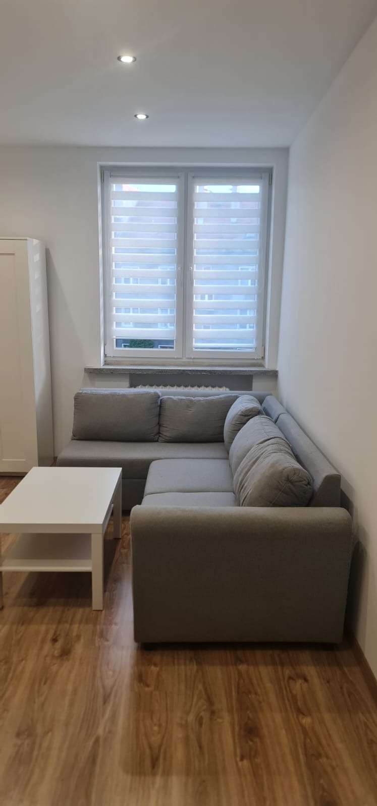 Jasne wnętrze po malowaniu: szary narożnik, białe ściany, stolik kawowy i okno z żaluzjami. Podłoga z paneli imitujących drewno, minimalistyczny styl.