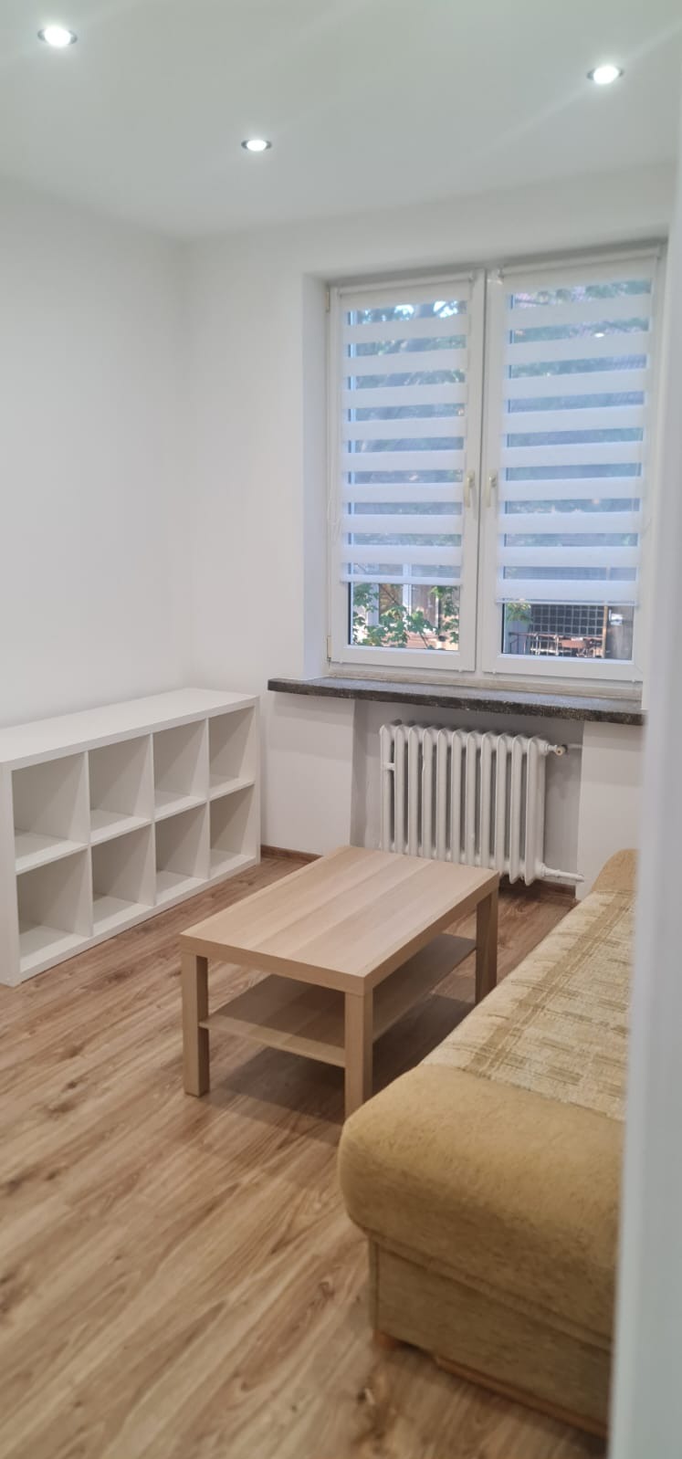 Jasne wnętrze pokoju po odświeżeniu ścian, z nową podłogą i meblami. Widoczne białe ściany, okno z roletami i grzejnik.
