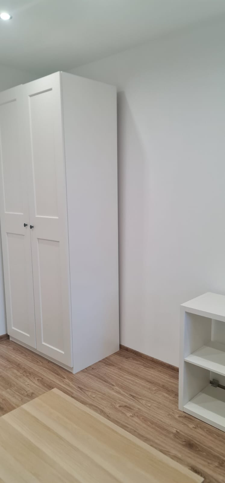 Wnętrze pokoju po remoncie: biała szafa z ciemnymi uchwytami, biała ściana, drewniana podłoga i biały regał. Minimalistyczny styl i świeżo pomalowane ściany.