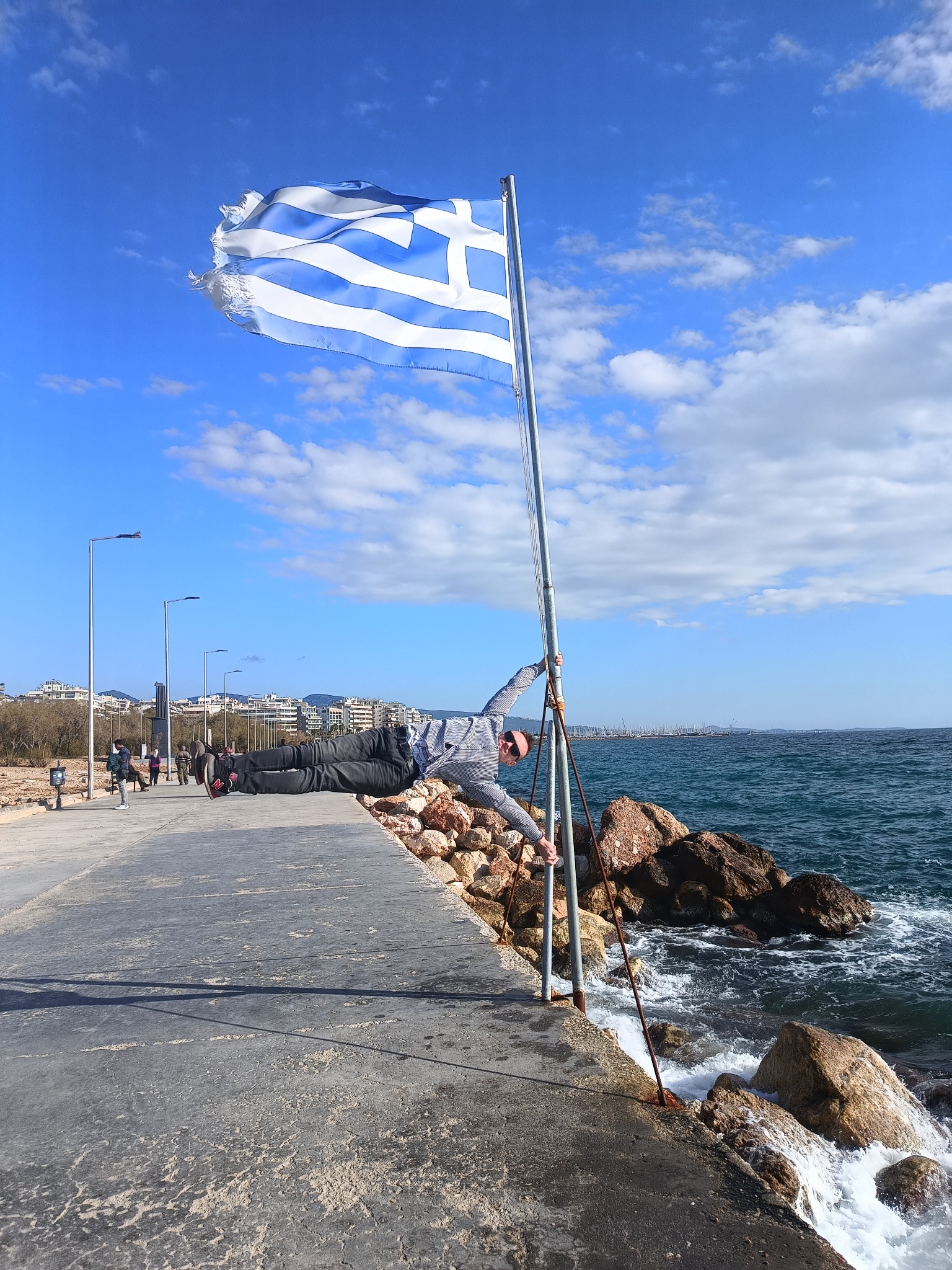 Mężczyzna w pozycji deski, opierający się o maszt z grecką flagą na tle morza i błękitnego nieba z chmurami.