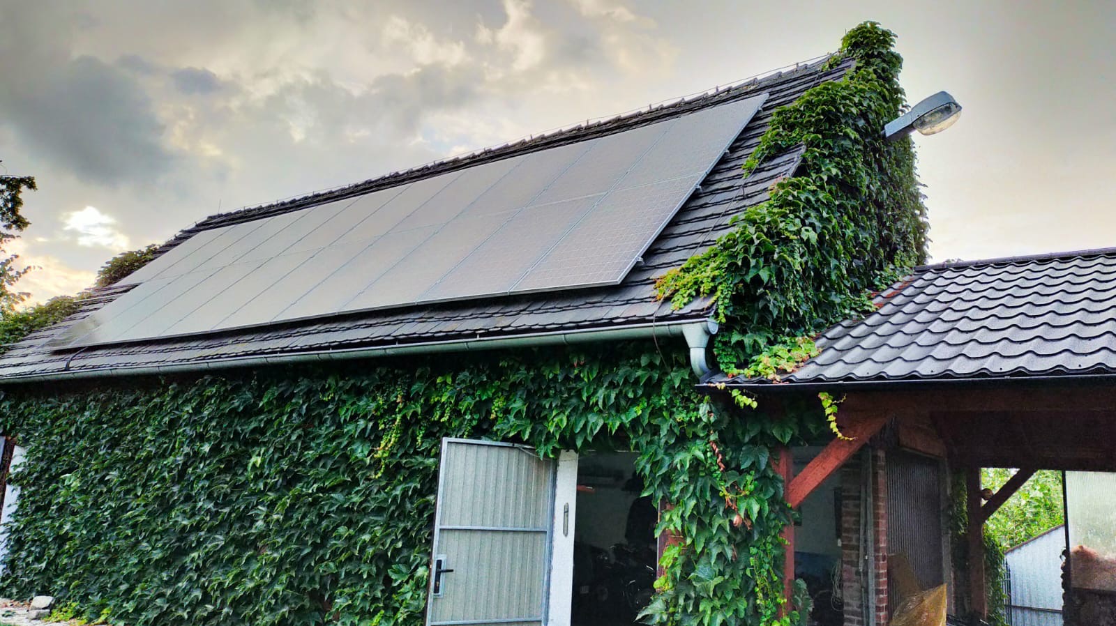 Panele słoneczne na dachu budynku obrośniętego bluszczem, z widocznym oświetleniem zewnętrznym i otwartymi drzwiami garażowymi. Nietypowe połączenie natury i technologii.