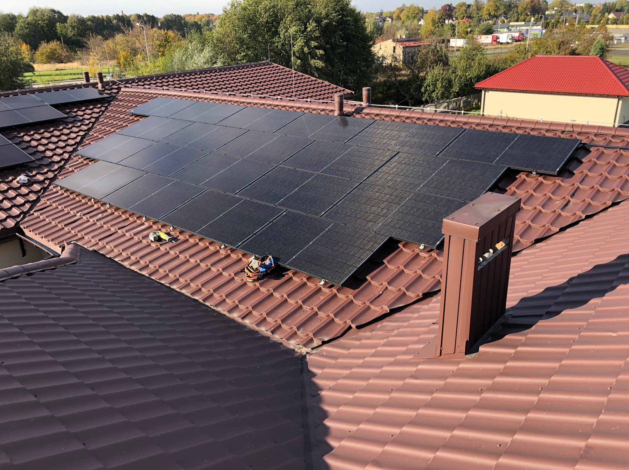 Panele fotowoltaiczne na dachu z brązowej dachówki, widok z góry. Częściowo zainstalowane, narzędzia obok. W tle zabudowa jednorodzinna i drzewa.