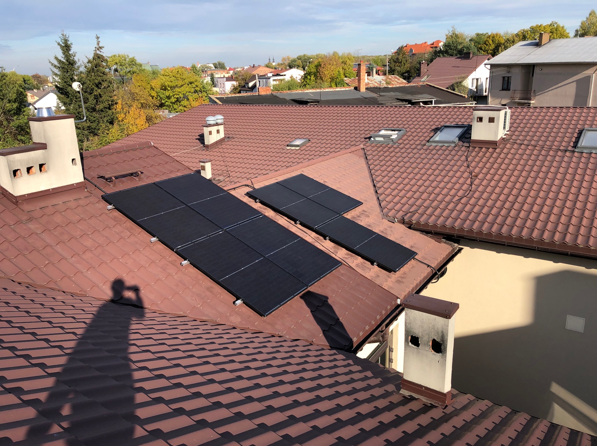Panele słoneczne na brązowym dachu domu z kominem i oknami dachowymi, cień fotografa widoczny na dachówce. Tło: zabudowa miejska i drzewa.