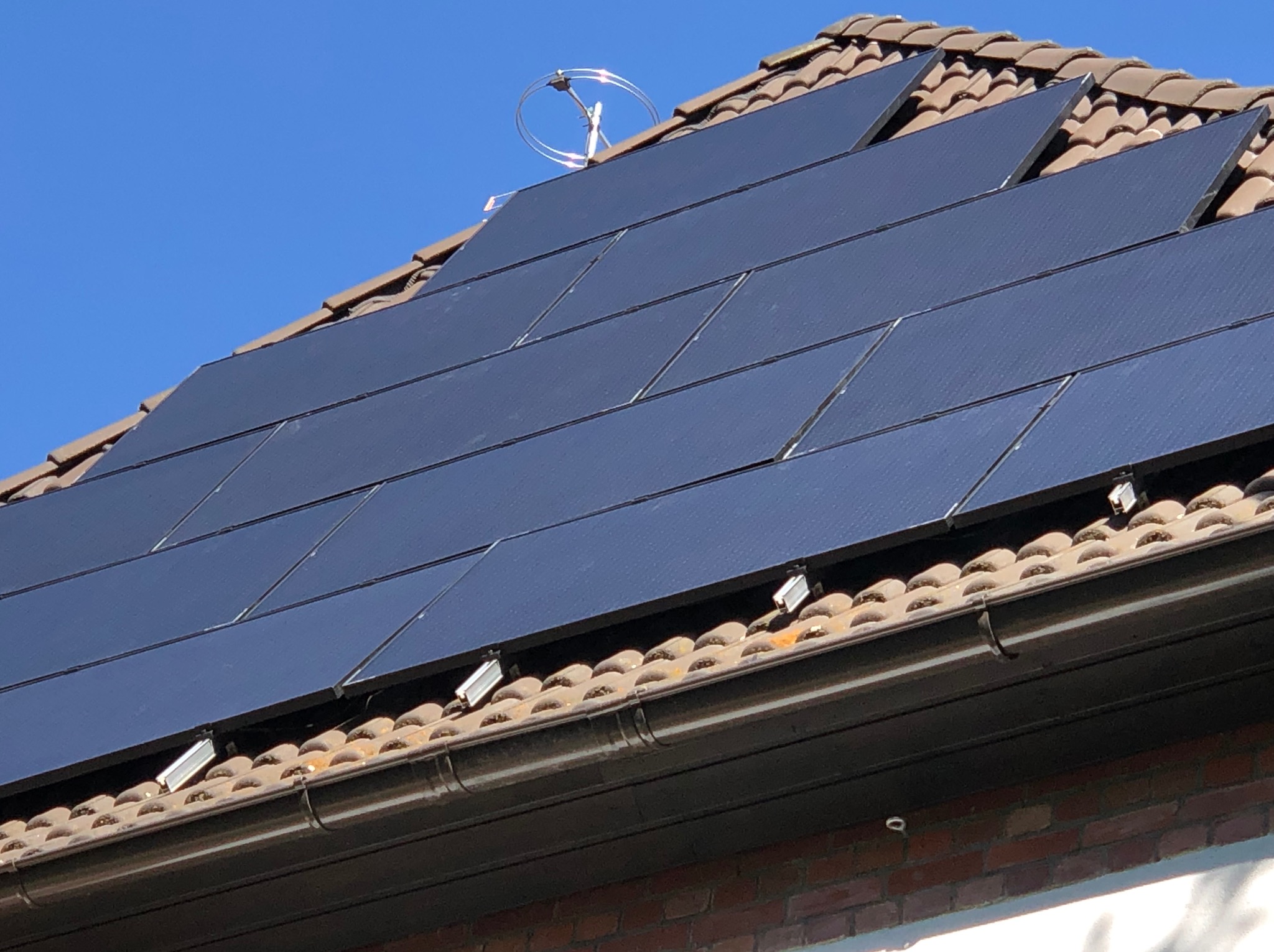 Panele słoneczne na dachu z brązowej dachówki, z widocznym uchwytem antenowym, na tle błękitnego nieba. Widoczne mocowania paneli.