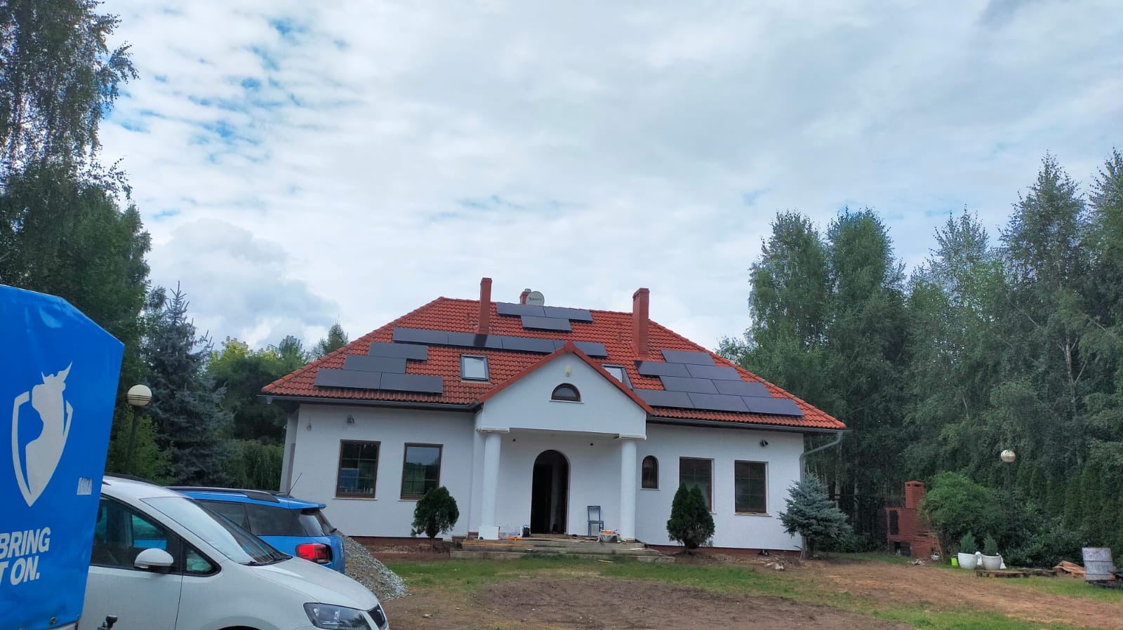 Dom jednorodzinny z czerwoną dachówką i panelami słonecznymi na dachu. Widoczny fragment samochodu dostawczego z logotypem firmy. Ujęcie w słoneczny dzień.