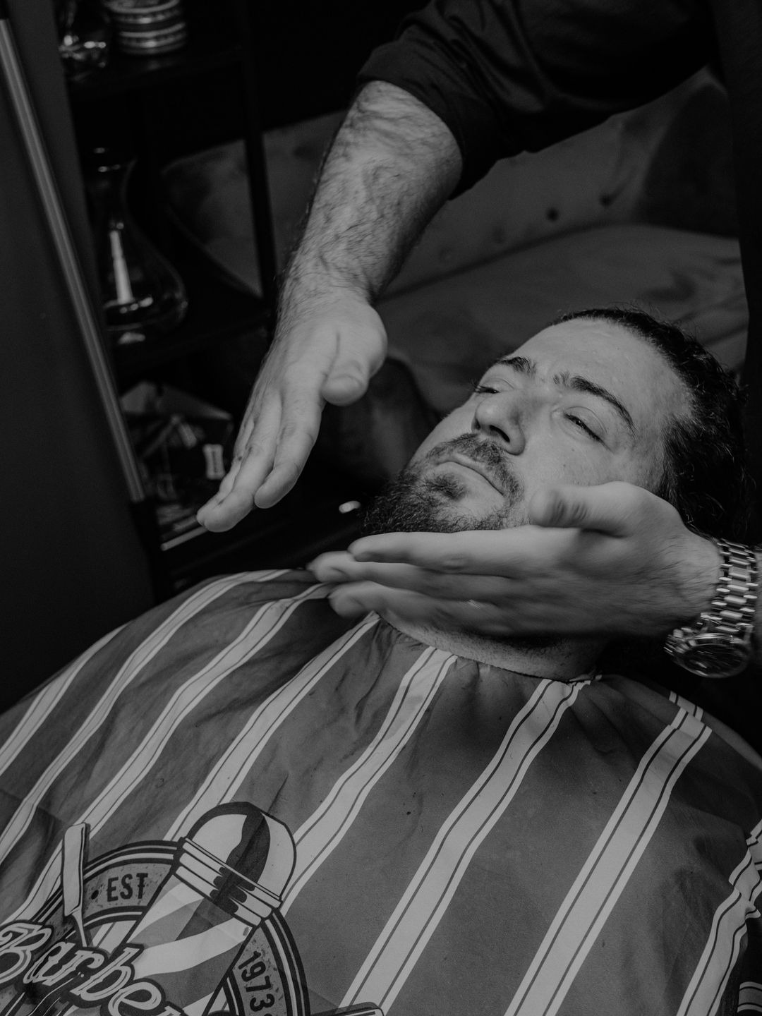 Masaż twarzy klienta w salonie, mężczyzna z brodą leży z zamkniętymi oczami, na nim peleryna w paski, widoczny zegarek na ręce masującego.
