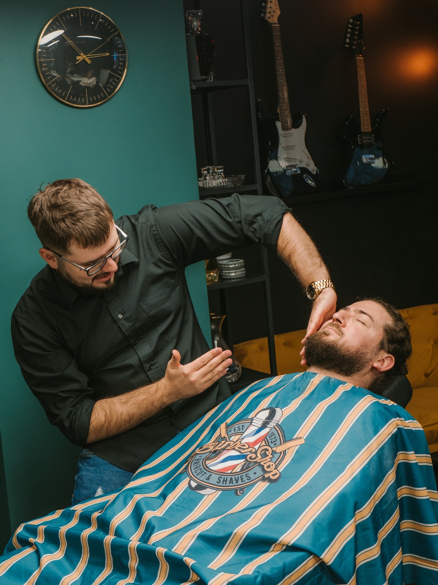 Mężczyzna z brodą leży na fotelu pod peleryną w niebieskie i żółte paski z logo Barber Shop, podczas gdy stylista w czarnej koszuli i okularach przygotowuje się do golenia jego brody. W tle...