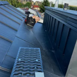 Nowoczesny, czarny dach z blachy na tle zabudowy miejskiej, widoczny fragment kratki wentylacyjnej i torba narzędziowa.