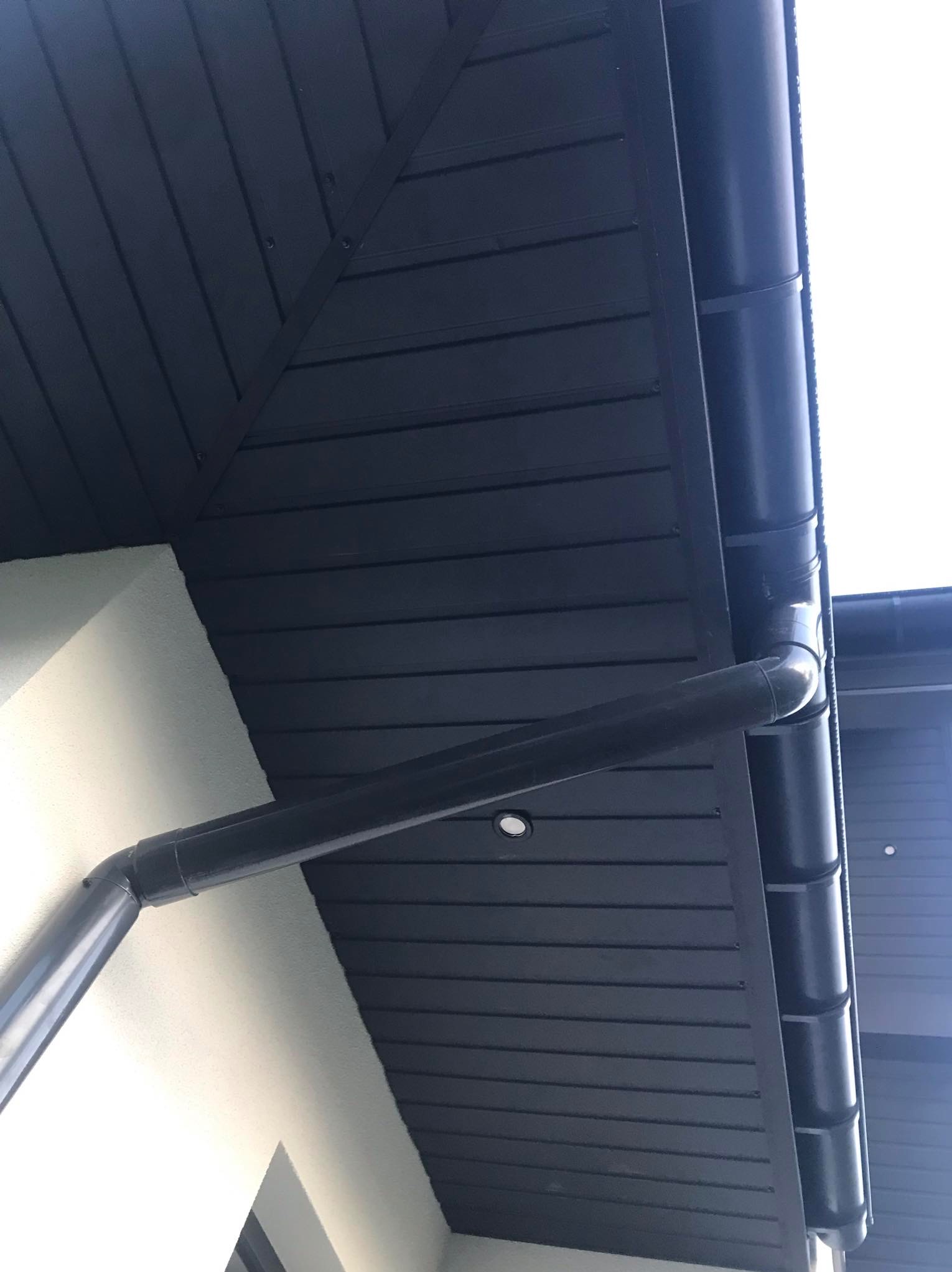 Elewacja domu z widocznym, ciemnym dachem z blachy trapezowej i systemem rynnowym w kolorze grafitowym, zamontowanym przy białej ścianie.