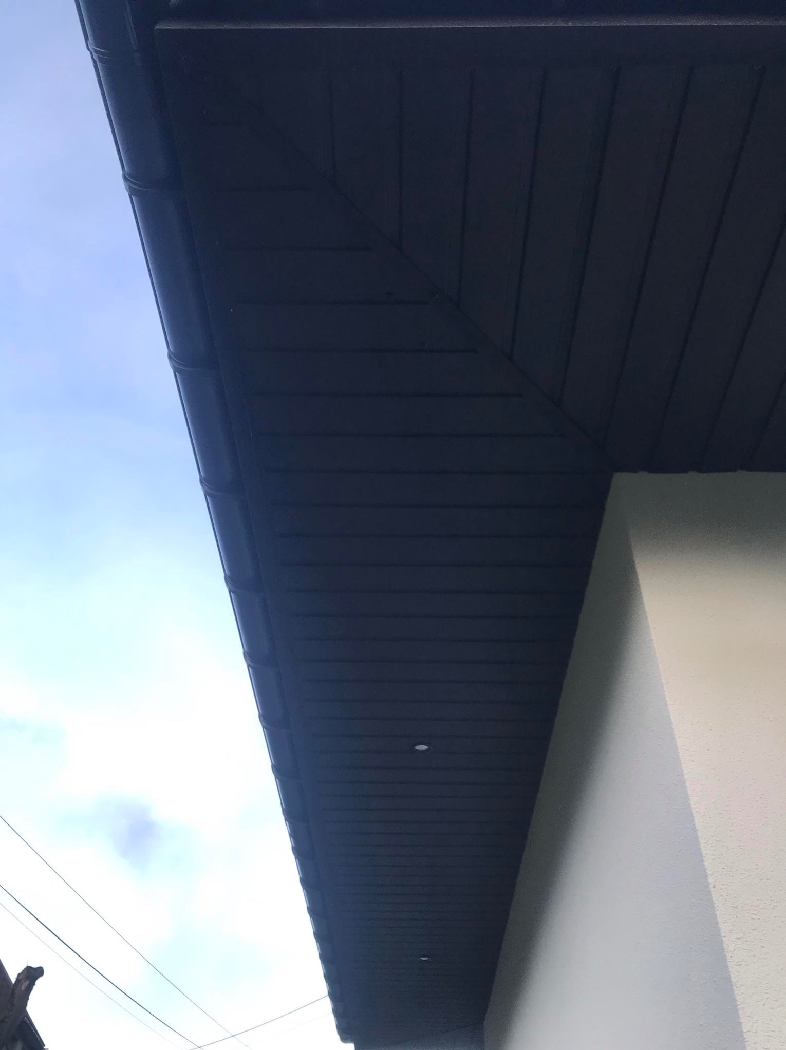 Nowoczesny, ciemny dach z podbitką w odcieniach szarości, widoczny z dołu pod kątem, z fragmentem białej ściany budynku i błękitnym niebem w tle.